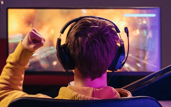 Человек в наушниках с микрофоном, воспроизводящим звук на компьютере, поднимает сжатый кулак перед монитором игры.