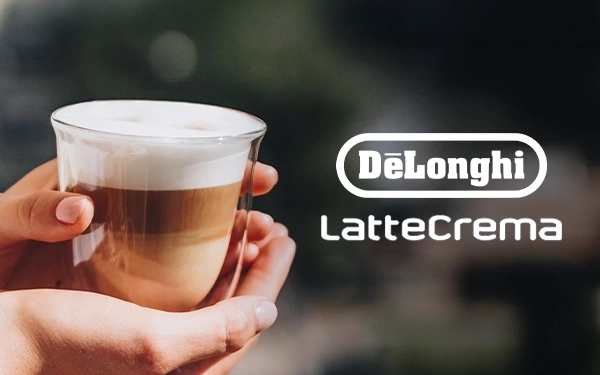 Руки тримають склянку пінистого лате поруч із логотипом DeLonghi LatteCrema на розмитому фоні
