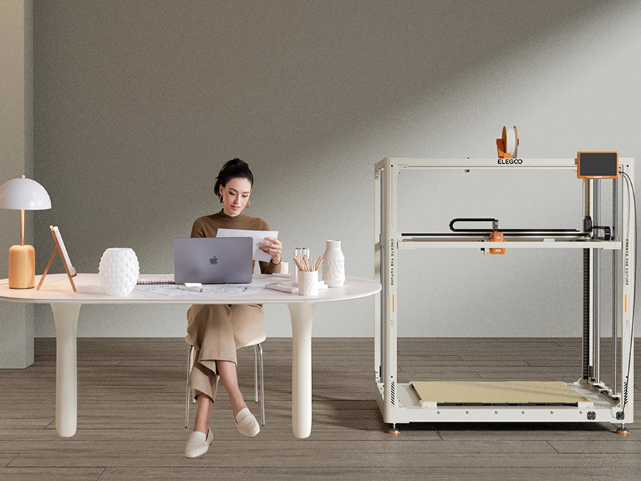 Жінка в бежевому вбранні працює на ноутбуці за білим столом поруч із білим 3D-принтером ELEGOO.
